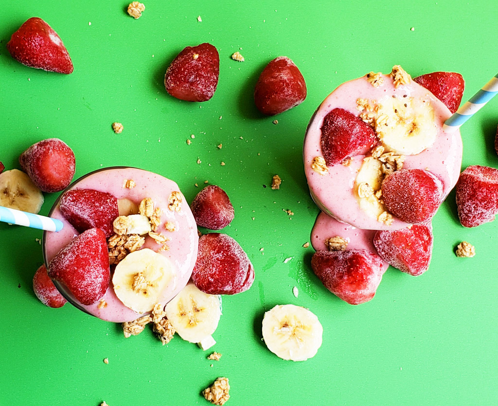 banana strawberry oat smoothie in a clear glass topped with frozen strawberries, bananas, and granola