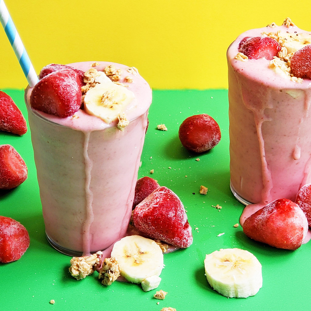 banana strawberry oat smoothie in a clear glass topped with frozen strawberries, bananas, and granola