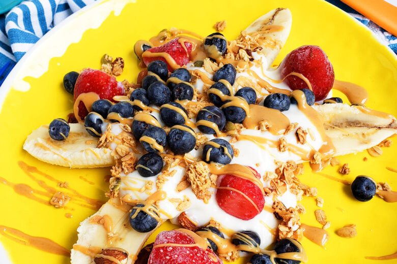 Healthy Breakfast Banana Split, banana sliced in half topped with Greek yogurt, blueberries, strawberries, granola, and peanut butter drizzle on a yellow plate