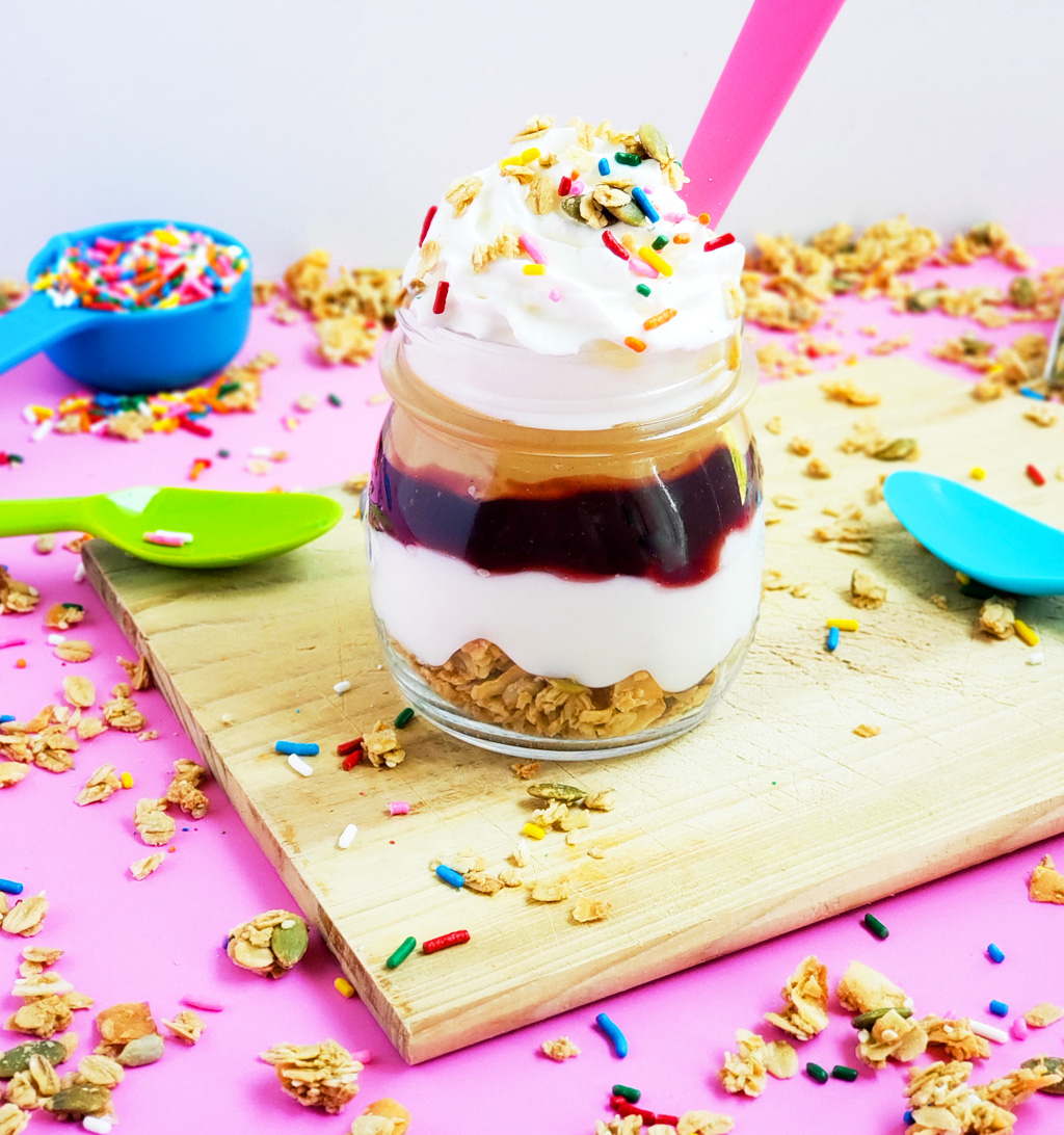 PB&J Greek Yogurt Parfait made with Bob's Red Mill Granola, peanut butter and jelly and sprinkles on a pink surface