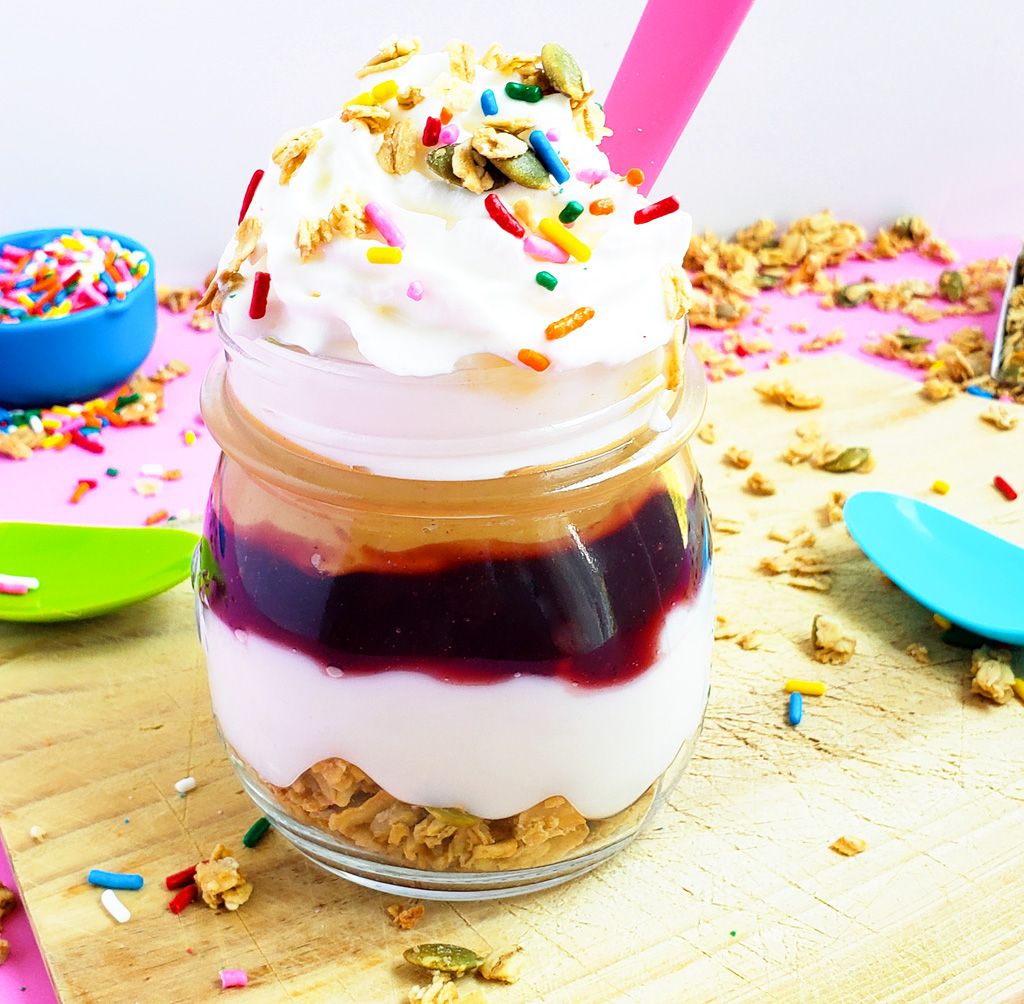 PB&J Greek Yogurt Parfait made with Bob's Red Mill Granola, peanut butter and jelly and sprinkles on a pink surface