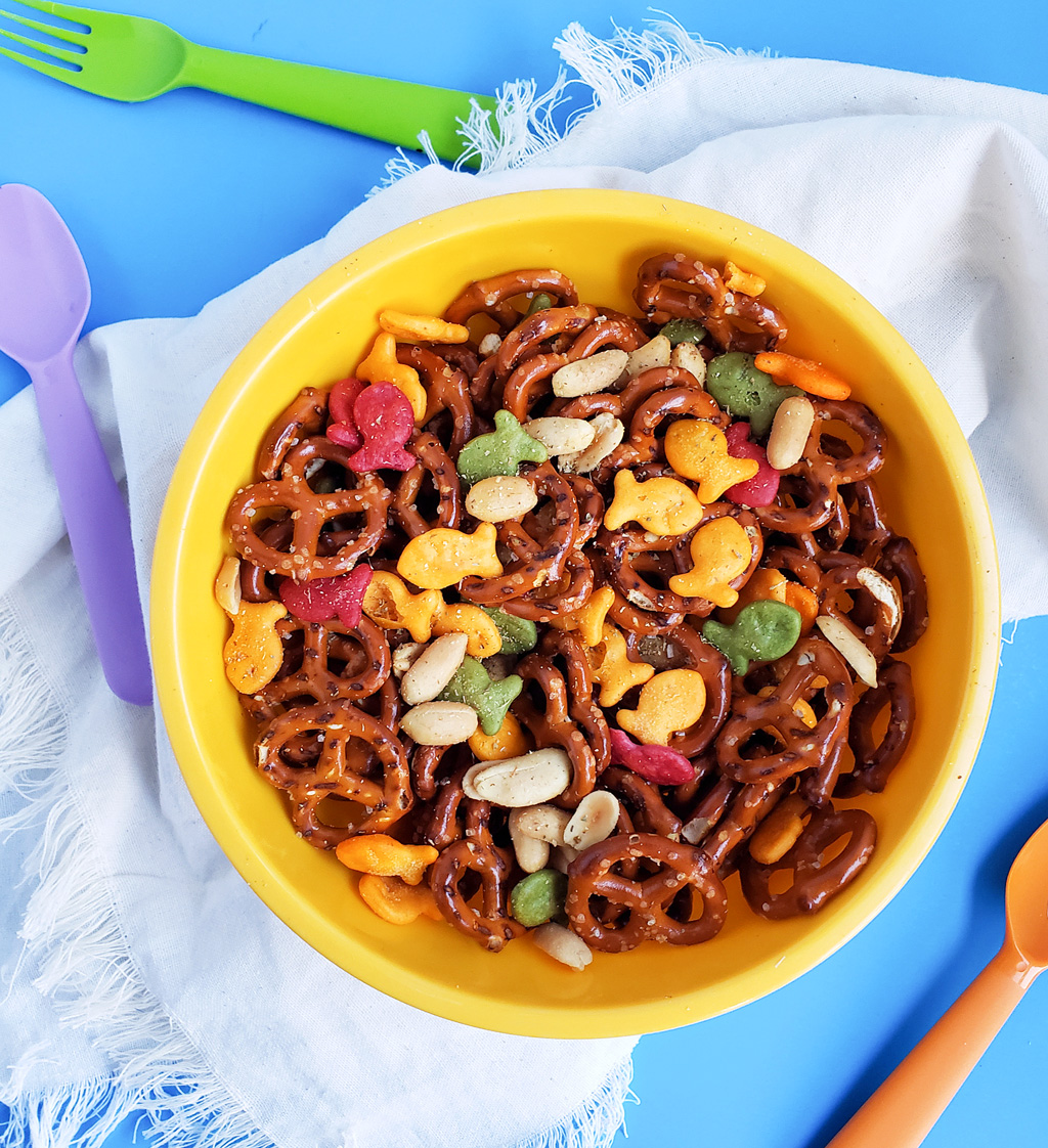 CJs Rockstar Trail Mix made with multi-colored goldfish crackers, pretzels, and peanuts in a yellow bowl on a blue surface