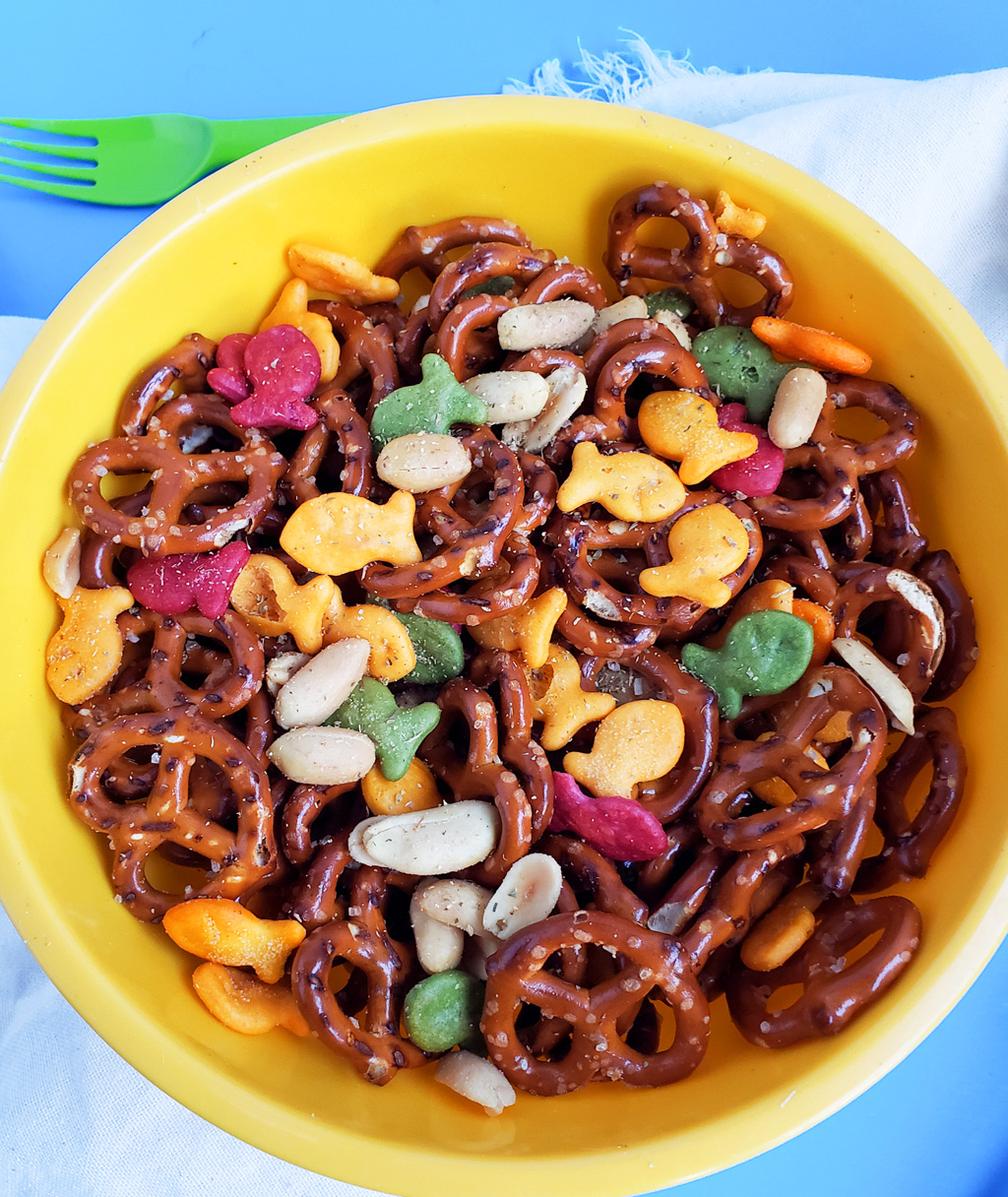 CJs Rockstar Trail Mix made with multi-colored goldfish crackers, pretzels, and peanuts in a yellow bowl on a blue surface