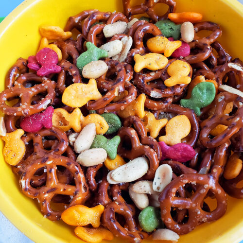 CJs Rockstar Trail Mix made with multi-colored goldfish crackers, pretzels, and peanuts in a yellow bowl on a blue surface