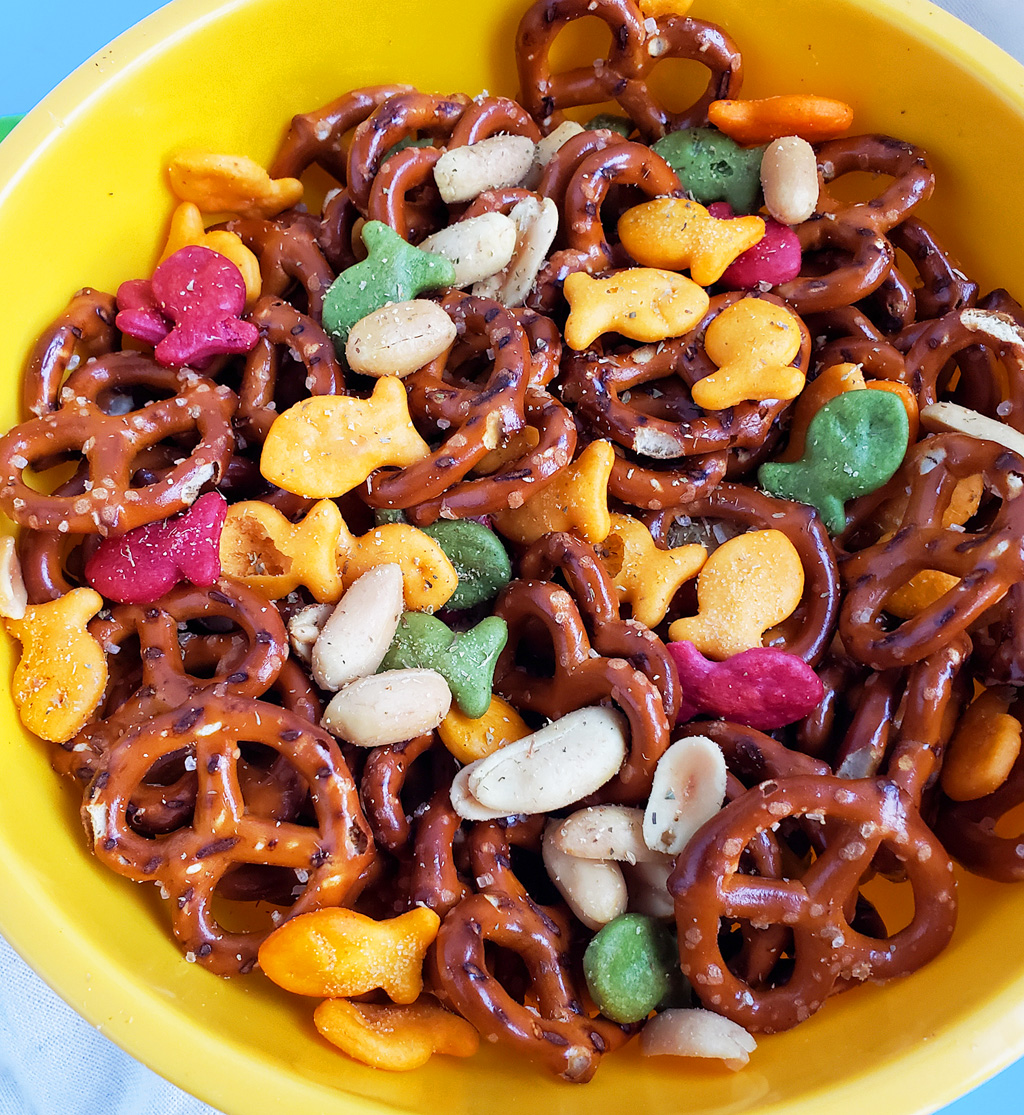 CJs Rockstar Trail Mix made with multi-colored goldfish crackers, pretzels, and peanuts in a yellow bowl on a blue surface