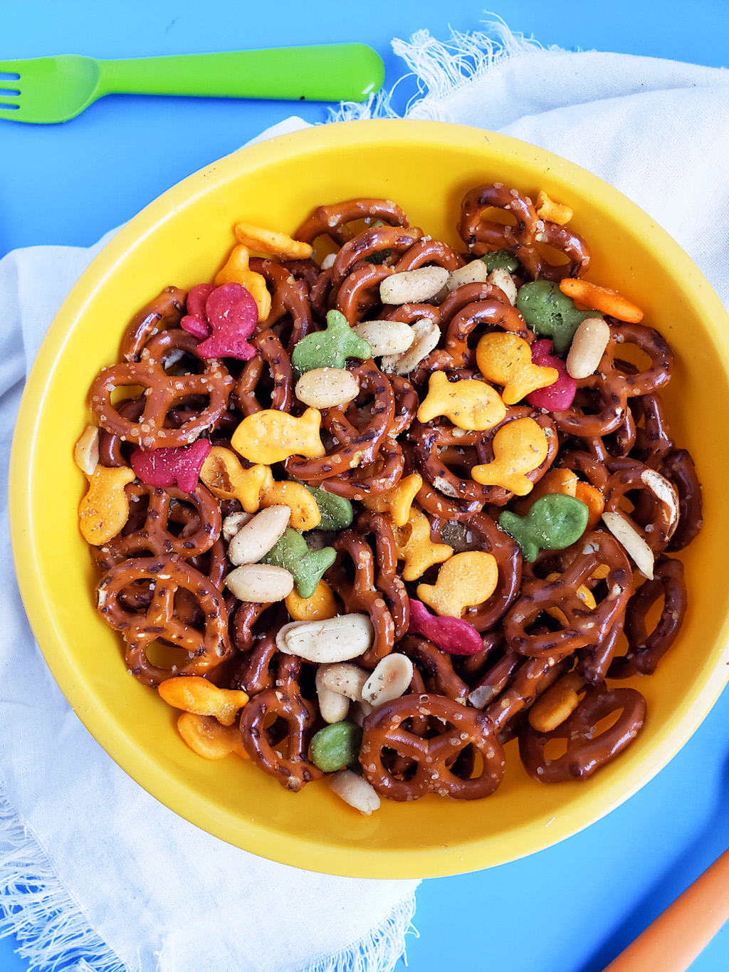 CJs Rockstar Trail Mix made with multi-colored goldfish crackers, pretzels, and peanuts in a yellow bowl on a blue surface