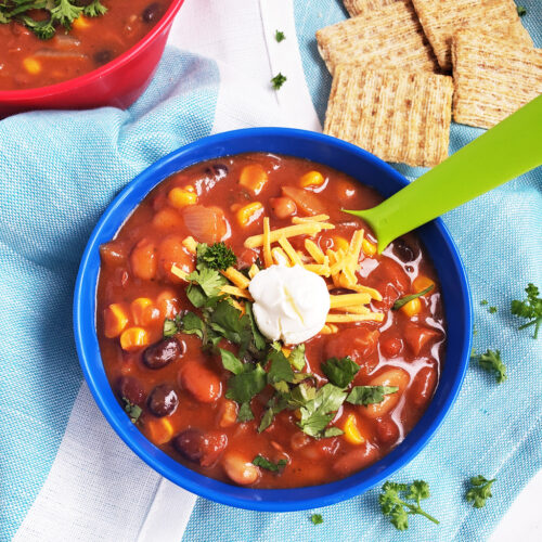 Simple Vegetarian Taco Soup, the perfect meatless meal, topped with green onion, cheese, and sour cream, served in a kid-friendly blue bowl with a green spoon. #tacosoup #vegetarianmeals #plantbasedkids #plantbasedmeals #plantbasedkidmeals