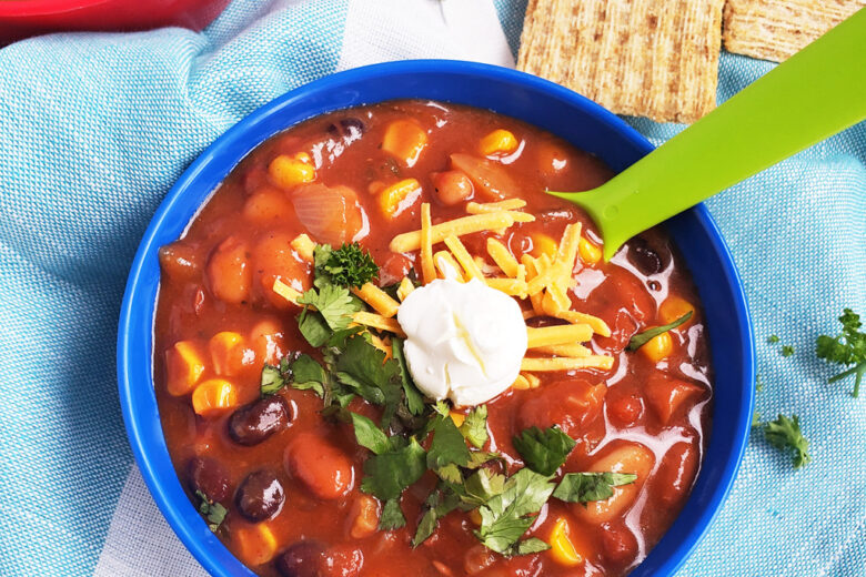 Simple Vegetarian Taco Soup, the perfect meatless meal, topped with green onion, cheese, and sour cream, served in a kid-friendly blue bowl with a green spoon. #tacosoup #vegetarianmeals #plantbasedkids #plantbasedmeals #plantbasedkidmeals