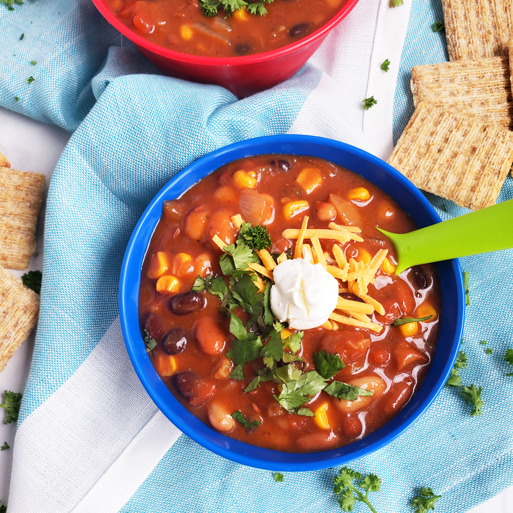 Simple Vegetarian Taco Soup, the perfect meatless meal, topped with green onion, cheese, and sour cream, served in a kid-friendly blue bowl with a green spoon. #tacosoup #vegetarianmeals #plantbasedkids #plantbasedmeals #plantbasedkidmeals