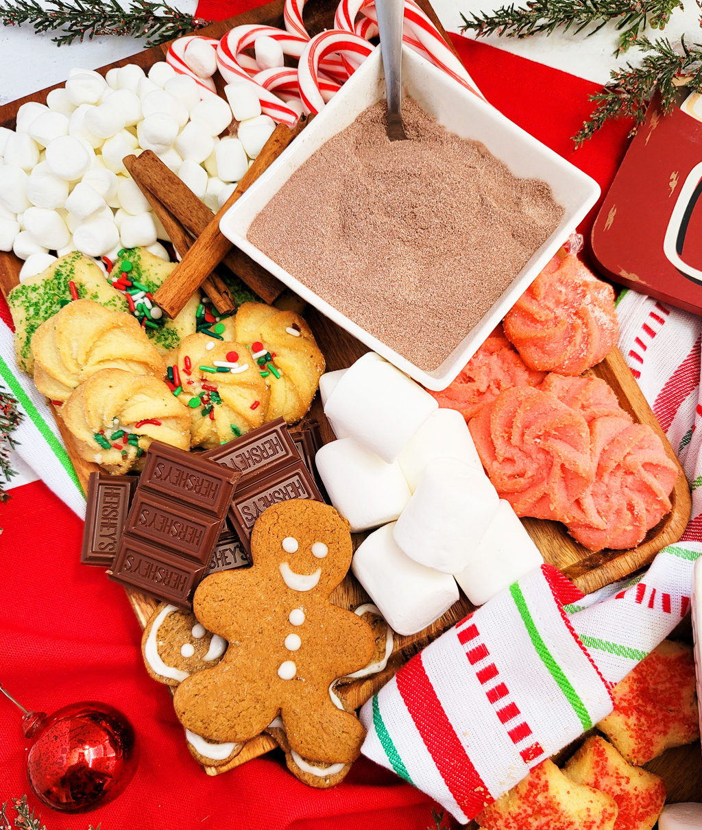 DIY Holiday Hot Cocoa Charcuterie Board on a wooden cutting board with gingerbread cookies, marshmallows, and hot chocolate mix. The perfect Christmas charcuterie board. www.littleeatsandthings.com 