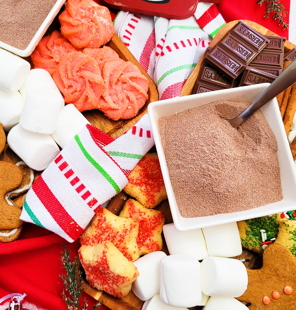 DIY Holiday Hot Cocoa Charcuterie Board on a wooden cutting board with gingerbread cookies, marshmallows, and hot chocolate mix. The perfect Christmas charcuterie board. www.littleeatsandthings.com 