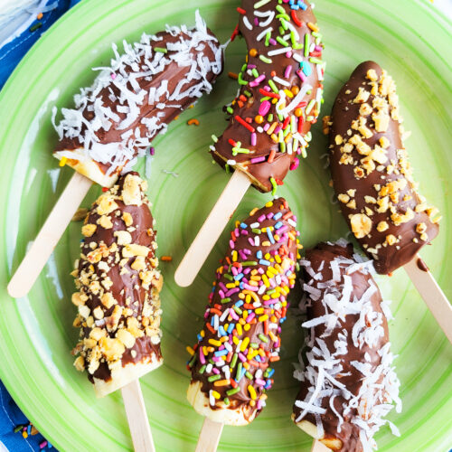 Frozen Chocolate Covered Bananas topped with peanuts, sprinkles, and coconut on a green plate. littleeatsandthings.com