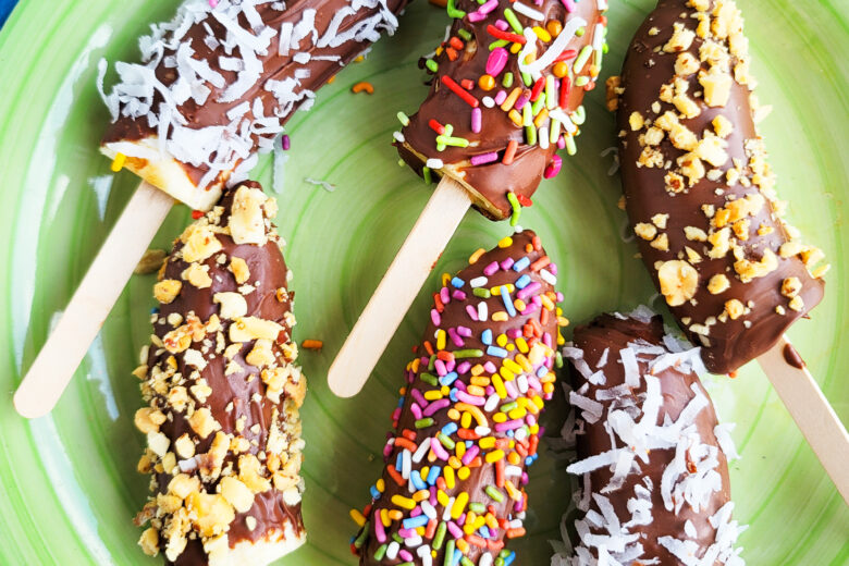 Frozen Chocolate Covered Bananas topped with peanuts, sprinkles, and coconut on a green plate. littleeatsandthings.com