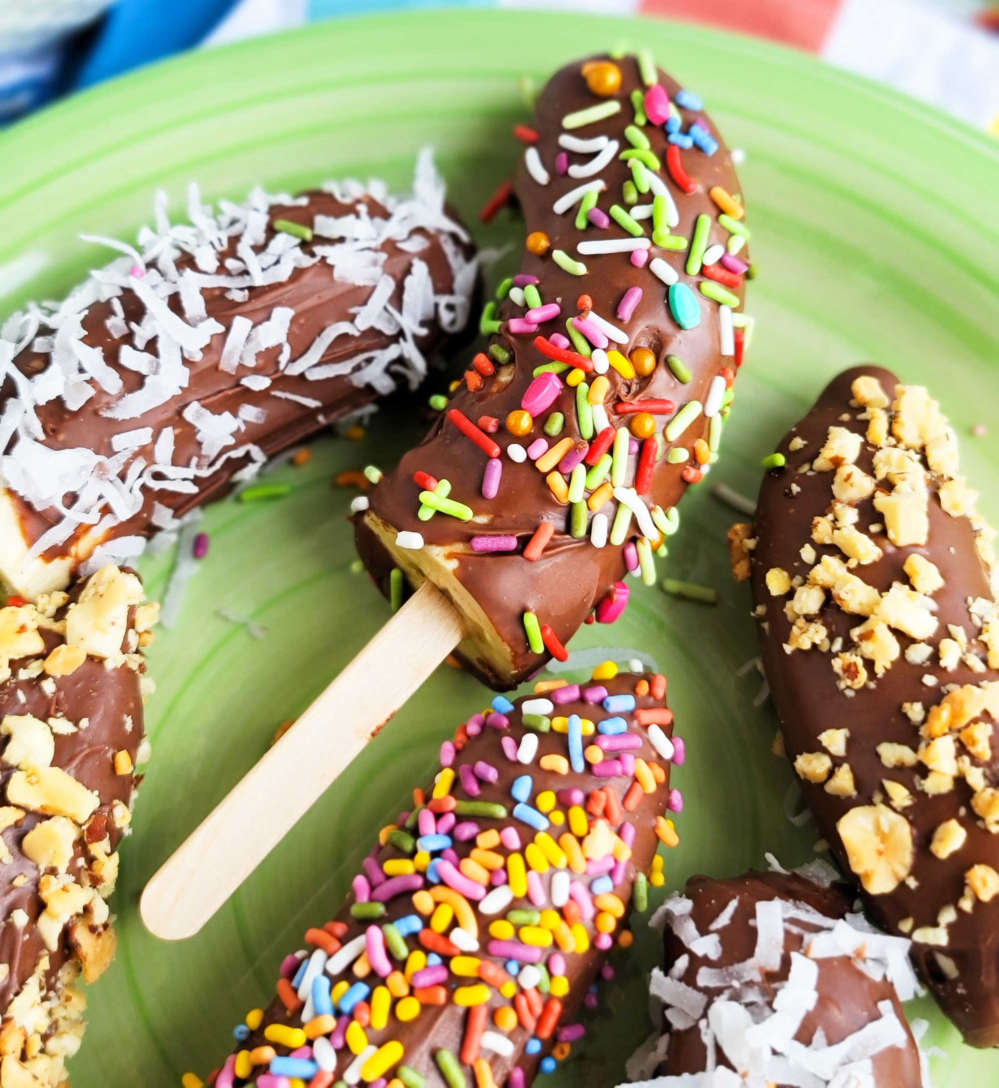 Frozen Chocolate Covered Bananas topped with peanuts, sprinkles, and coconut on a green plate. littleeatsandthings.com