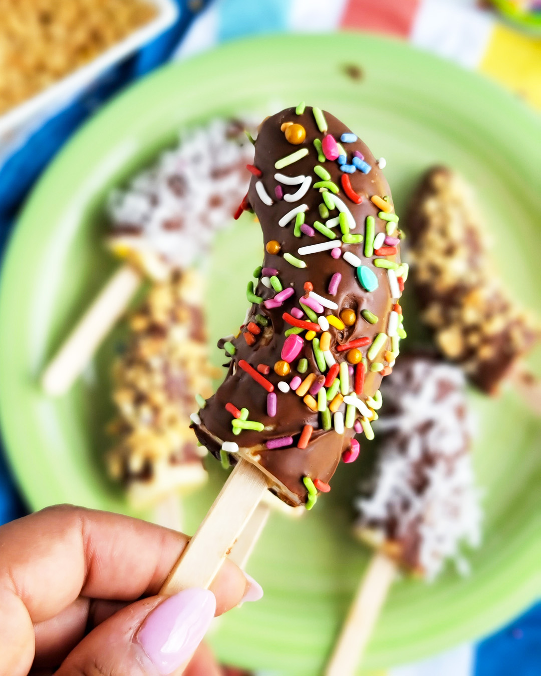 Frozen Chocolate Covered Bananas topped with peanuts, sprinkles, and coconut on a green plate. littleeatsandthings.com