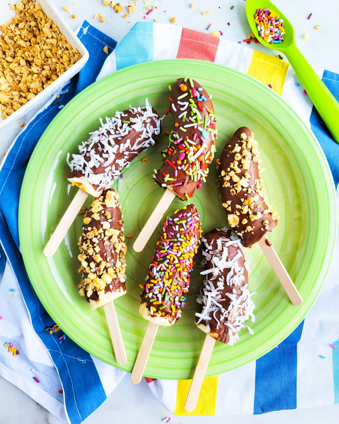 Frozen Chocolate Covered Bananas topped with peanuts, sprinkles, and coconut on a green plate. beautifuleatsandthings.com