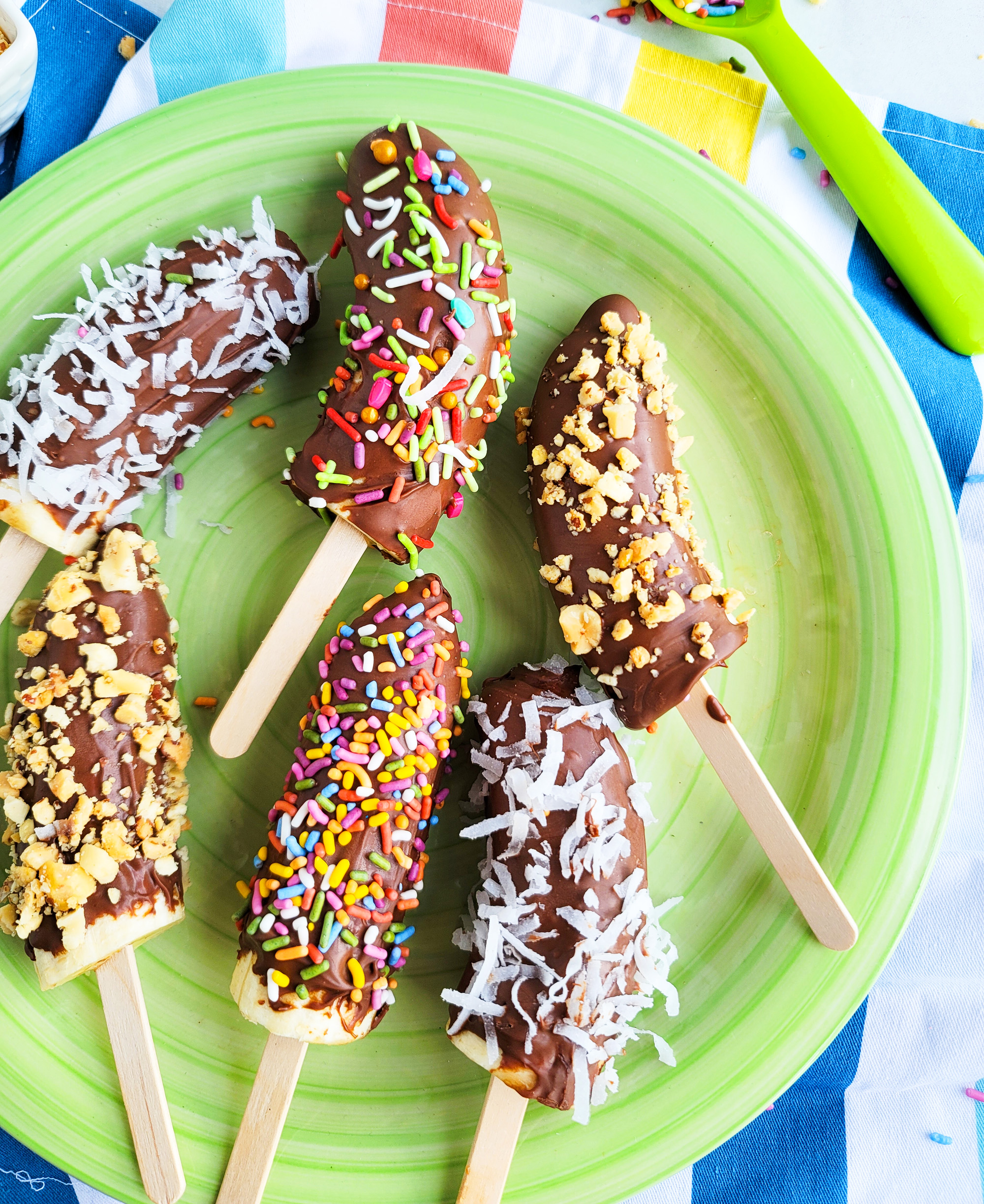 Frozen Chocolate Covered Bananas topped with peanuts, sprinkles, and coconut on a green plate. littleeatsandthings.com