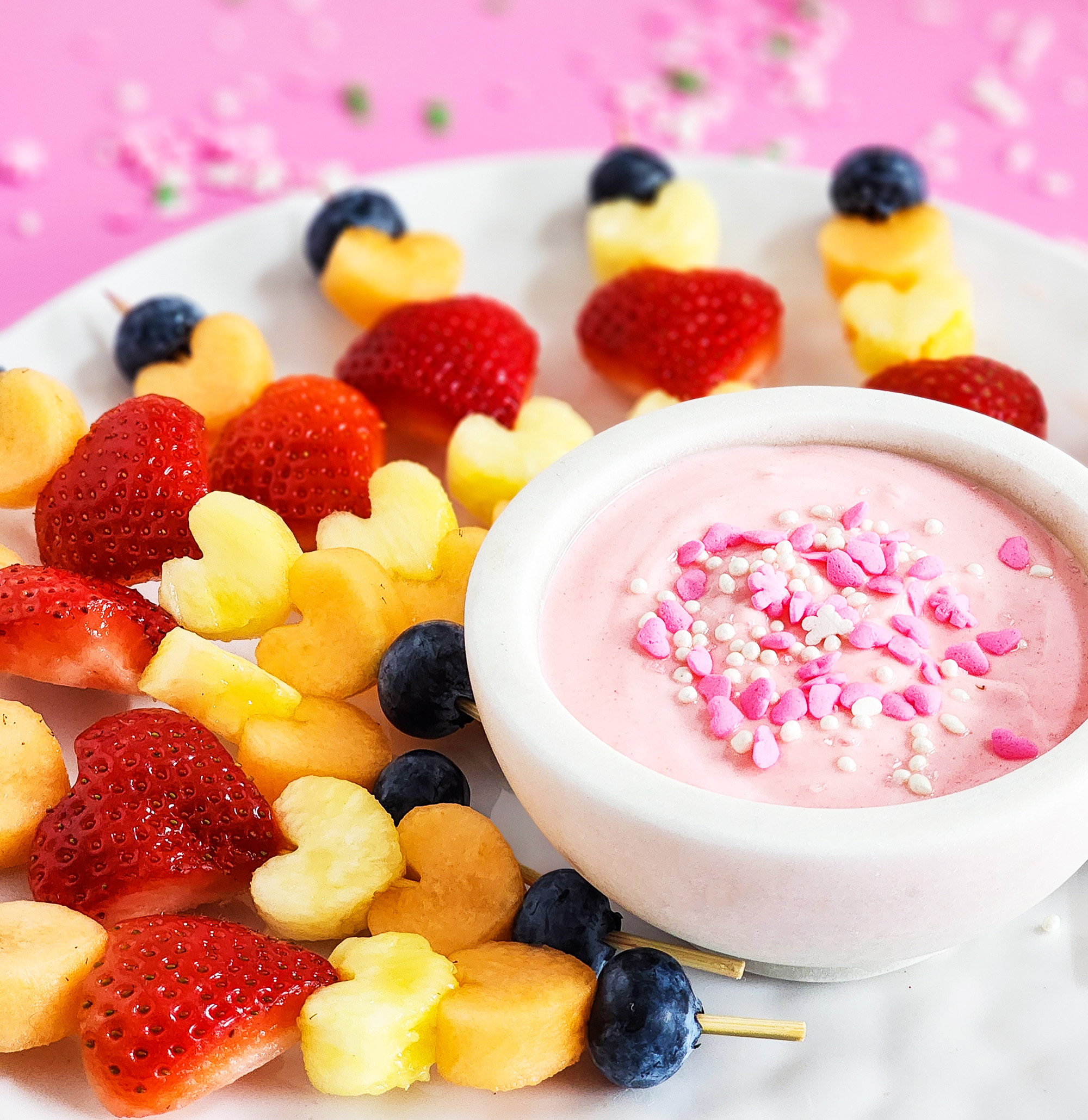 heart-shaped fruit skewers on a white plate on a pink surface for Valentine's Day littleeatsandthings.com